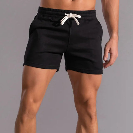 Black short-length shorts with a white drawstring and side pockets, worn by a model against a grey backdrop, front view. 1