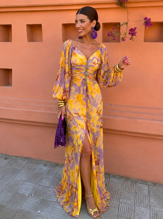 Smiling model in the yellow and lilac pleated maxi dress with V-neck, puffed sleeves, crystal trim and front slit, holding a purple clutch. 1