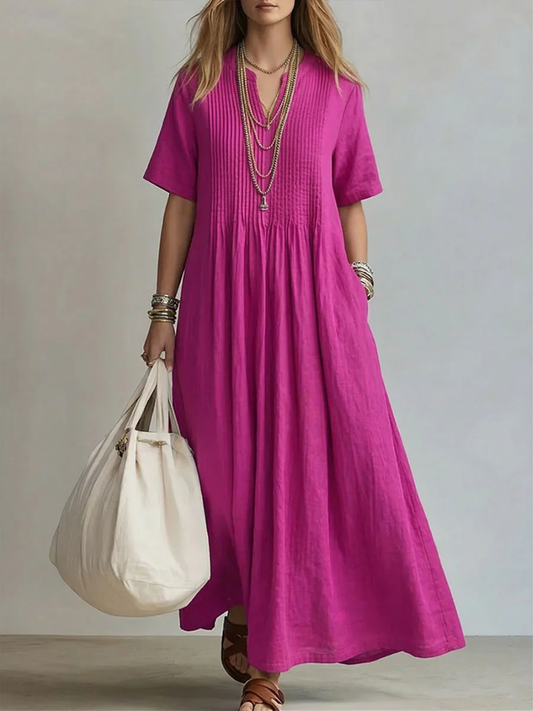 Model wearing the short-sleeved V-neck dress in magenta pink, with pintucked bodice, layered gold necklaces and a cream tote bag. 1