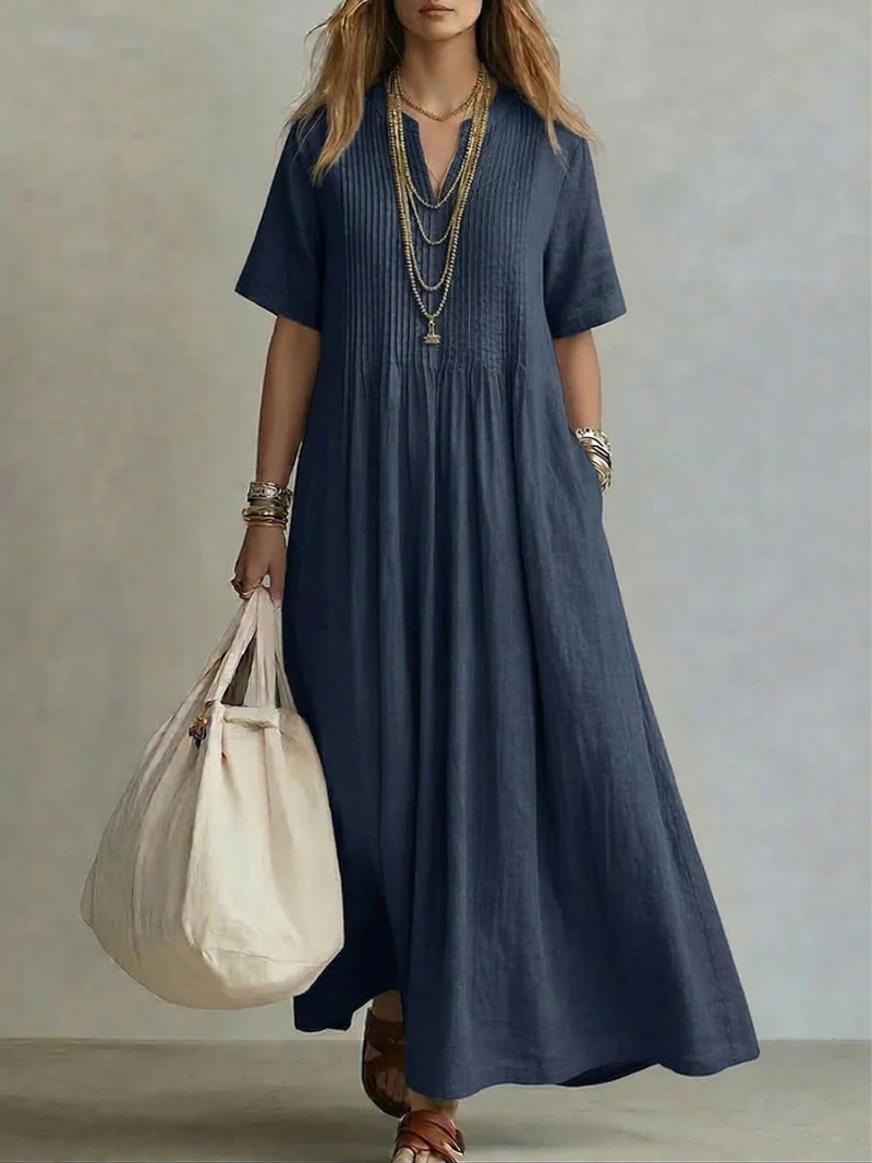 Model wearing the short-sleeved V-neck dress in deep navy blue, with pintucked bodice, layered gold necklaces and a cream tote bag. 5