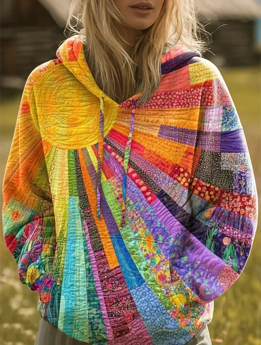 Woman with long blonde hair wearing the hoodie outdoors, its quilted patchwork of bright sun rays and floral panels visible, hands in pockets. 1