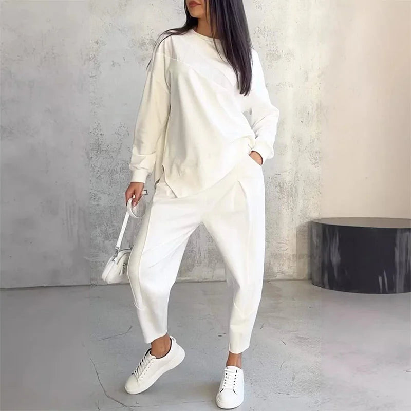 Model in the white set with one hand in a pocket, holding a small white bag, with white trainers, against a concrete wall. 1