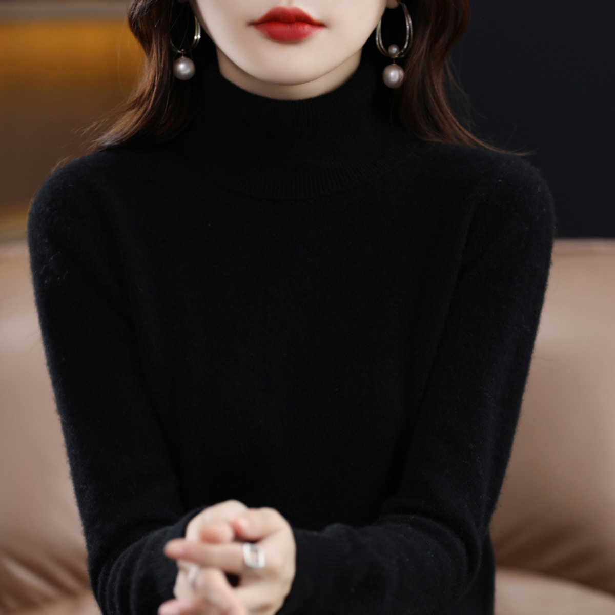 Close-up of a black roll neck jumper worn by a model, with hands clasped and pearl earrings visible. 2
