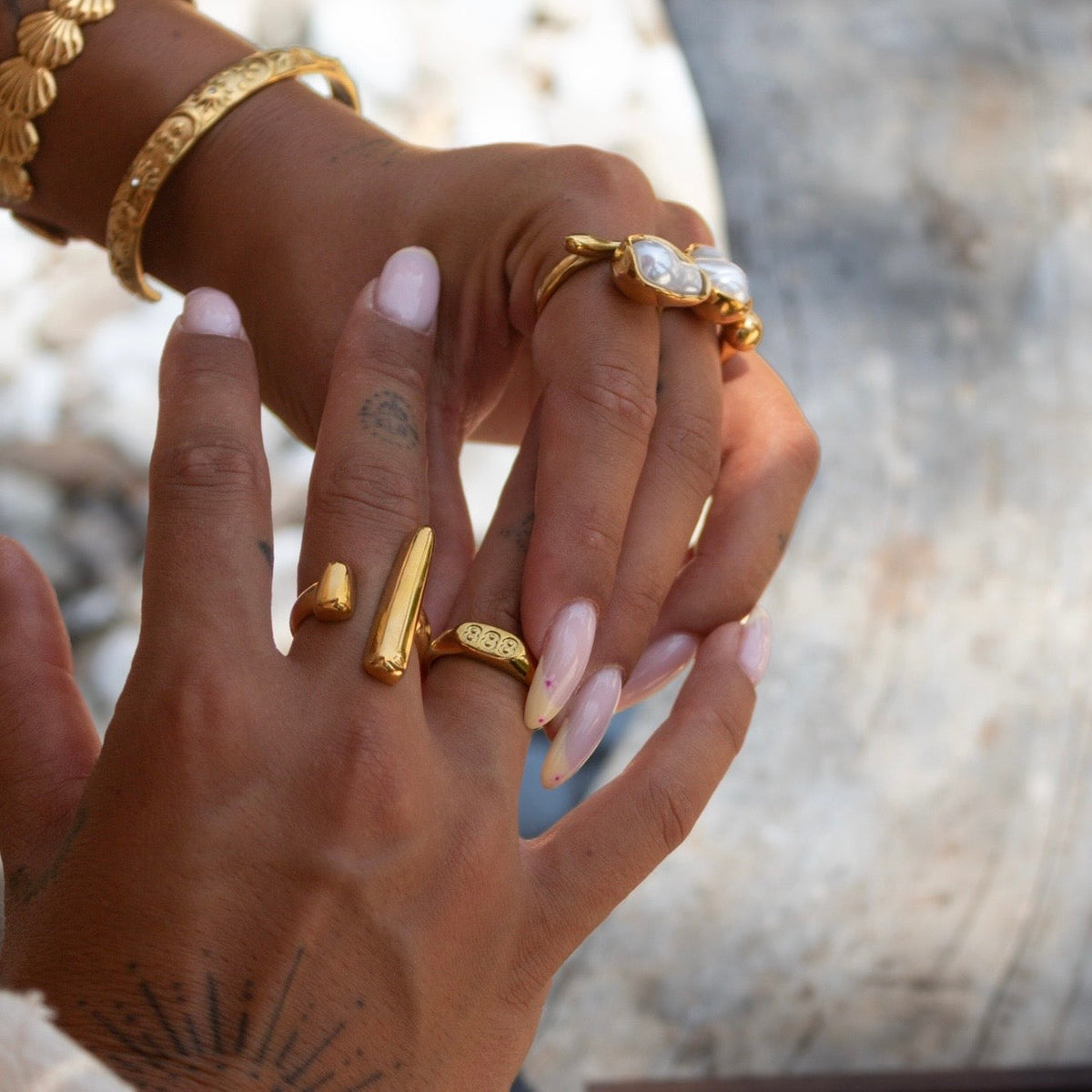 Hand wearing the open bar ring on one finger beside a number-engraved ring, with gold bangles and pearl rings behind. 5