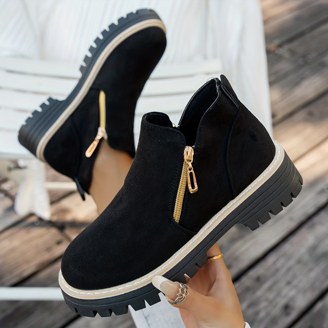 Black suede-look ankle boots with gold side zips and chunky treaded soles, worn by a model seated on a decking. 1