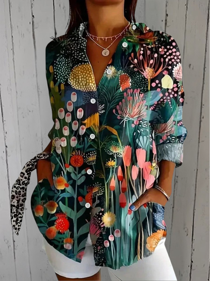 Model wearing the shirt in a dark green garden print of stylised flowers, tulips and dotted blooms, hands in pockets. 3
