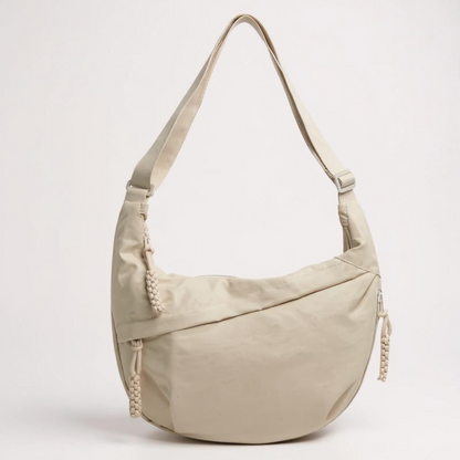 Beige crescent-shaped shoulder bag on a plain light background, with a looped adjustable strap and braided cord zip pulls at the sides. 1