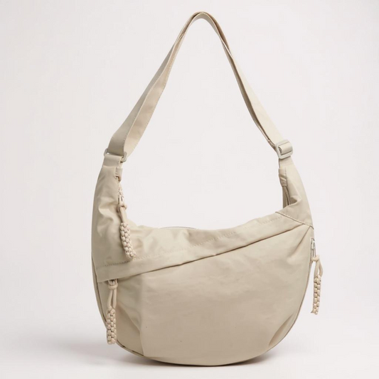 Beige crescent-shaped shoulder bag on a plain light background, with a looped adjustable strap and braided cord zip pulls at the sides. 1