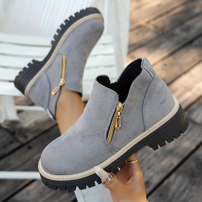 Grey suede-look ankle boots with gold side zips and chunky black treaded soles, worn by a model on wooden decking. 2