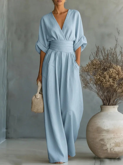 Pale blue version of the flowy wide-leg jumpsuit with rolled sleeves, worn by a model carrying a woven handbag in a studio. 4