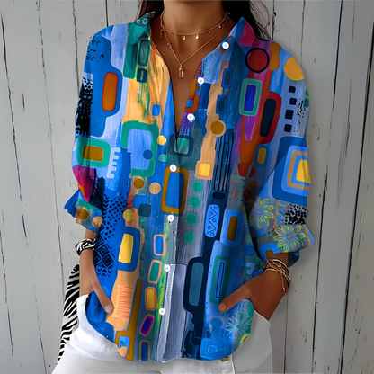 Model wearing the loose button-up blouse in a blue abstract print with orange and green accents, hands in the pockets of white trousers. 1