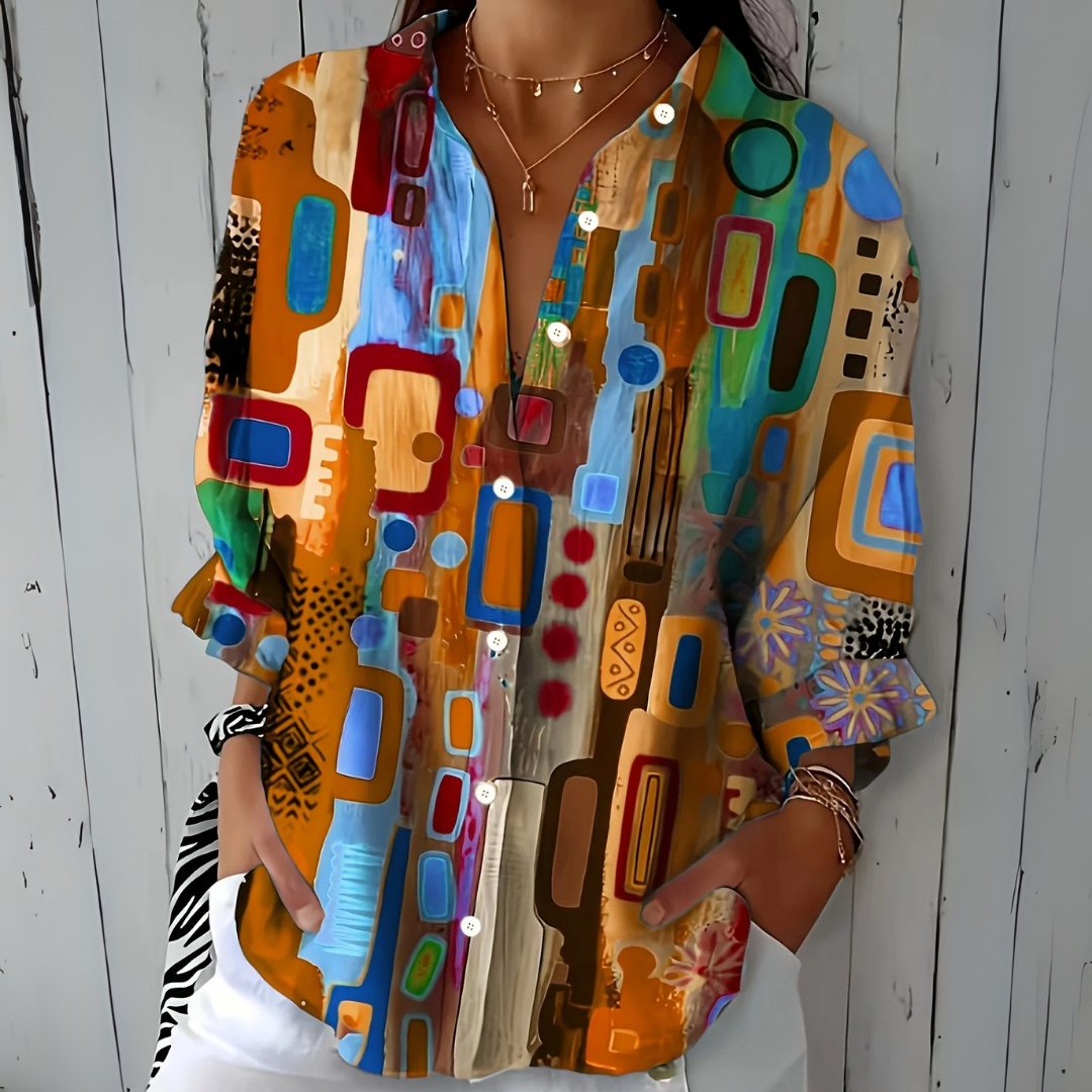 Model wearing the blouse in an orange and tan colourway with blue and red abstract shapes, styled with white trousers and layered necklaces. 3