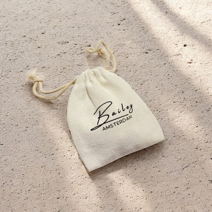 Cream drawstring cotton pouch with black printed lettering, lying on a sandy textured surface in dappled sunlight. 5
