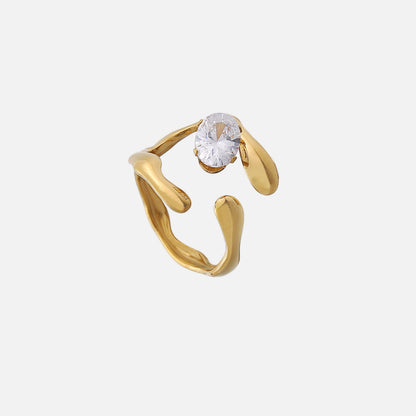 Gold-tone open ring with wavy, molten-looking band ends and a clear oval stone, shown from above on a plain light background. 1