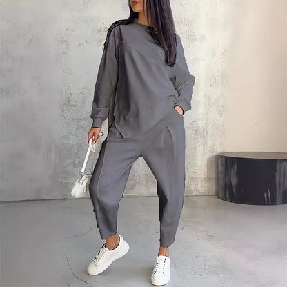 Model wearing the grey set, hand in pocket, with a small white bag and white trainers against a concrete wall. 3