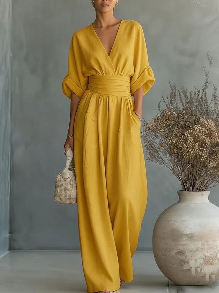 Mustard yellow version of the wide-leg jumpsuit with a wrap V-neck and wide waistband, worn in a studio next to a vase. 5
