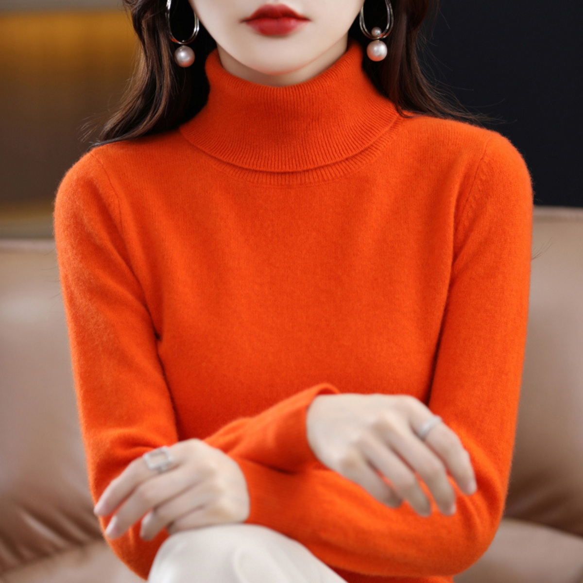 Model in the roll neck jumper in bright orange, hands crossed in front, with cream trousers. 10