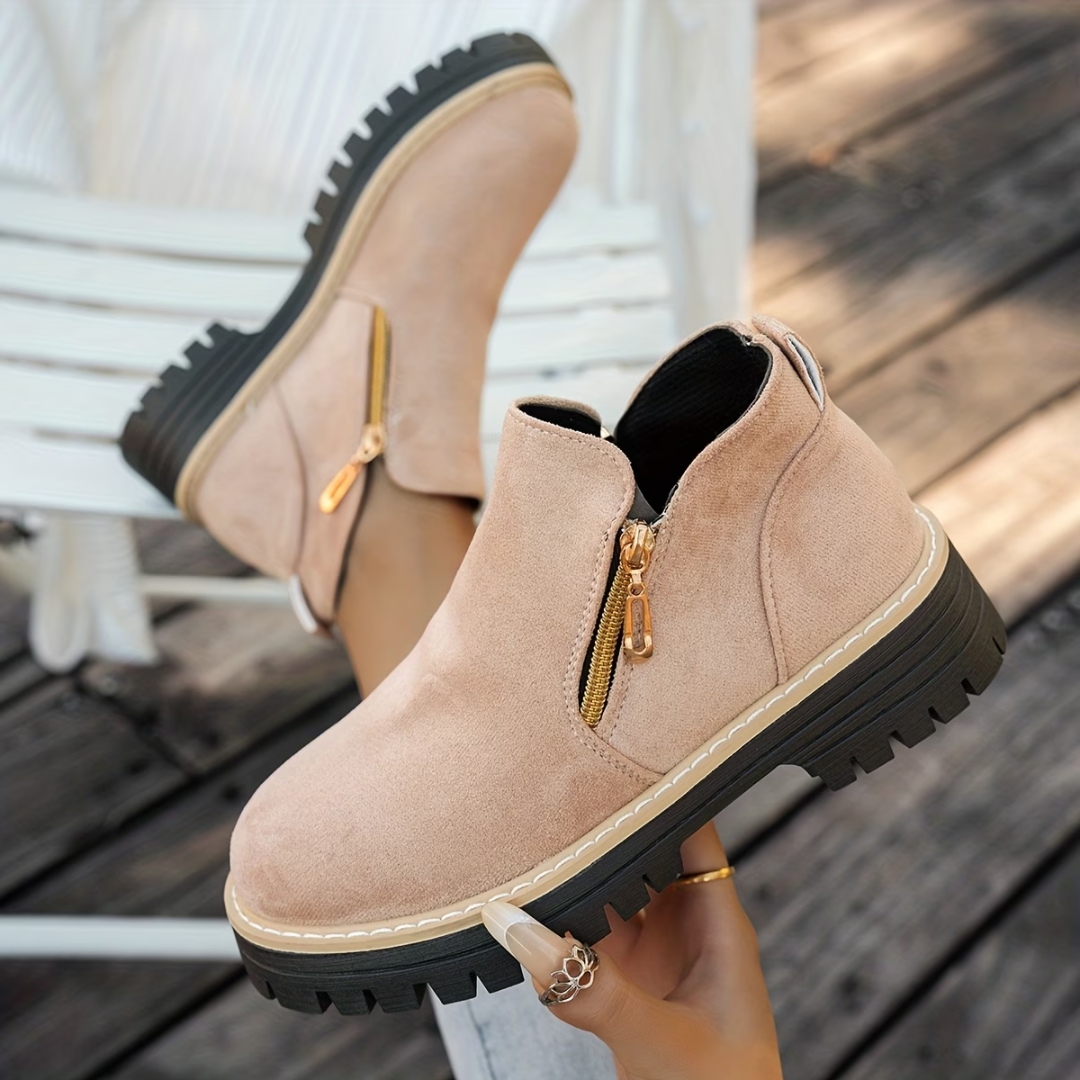Blush pink suede-look ankle boots with gold side zips and chunky treaded soles, worn by a model on wooden decking. 5