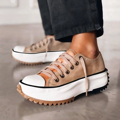 Side view of beige lace-up trainers with a chunky white platform sole and zigzag gum tread, worn with dark trousers. 1