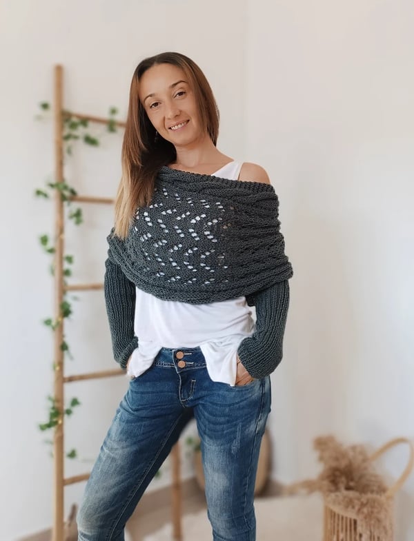 Dark grey lace-knit wrap with sleeves worn slightly off one shoulder, model smiling with hands in jeans pockets. 2