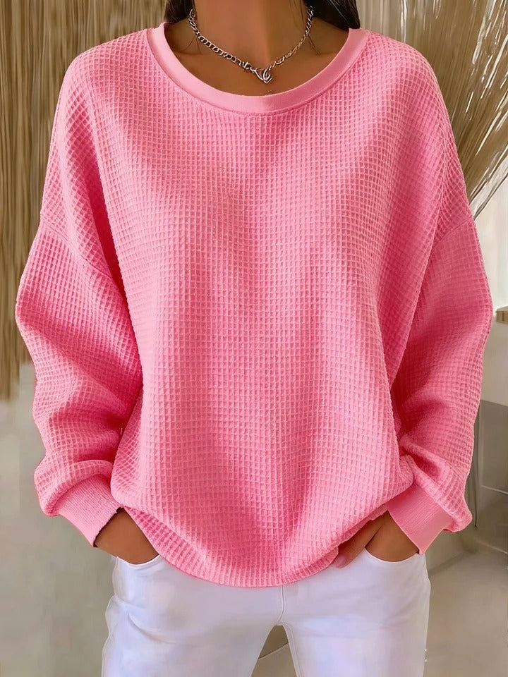 Model wearing the waffle-knit jumper in soft pink, with ribbed cuffs and hem, styled with white trousers and a necklace. 10