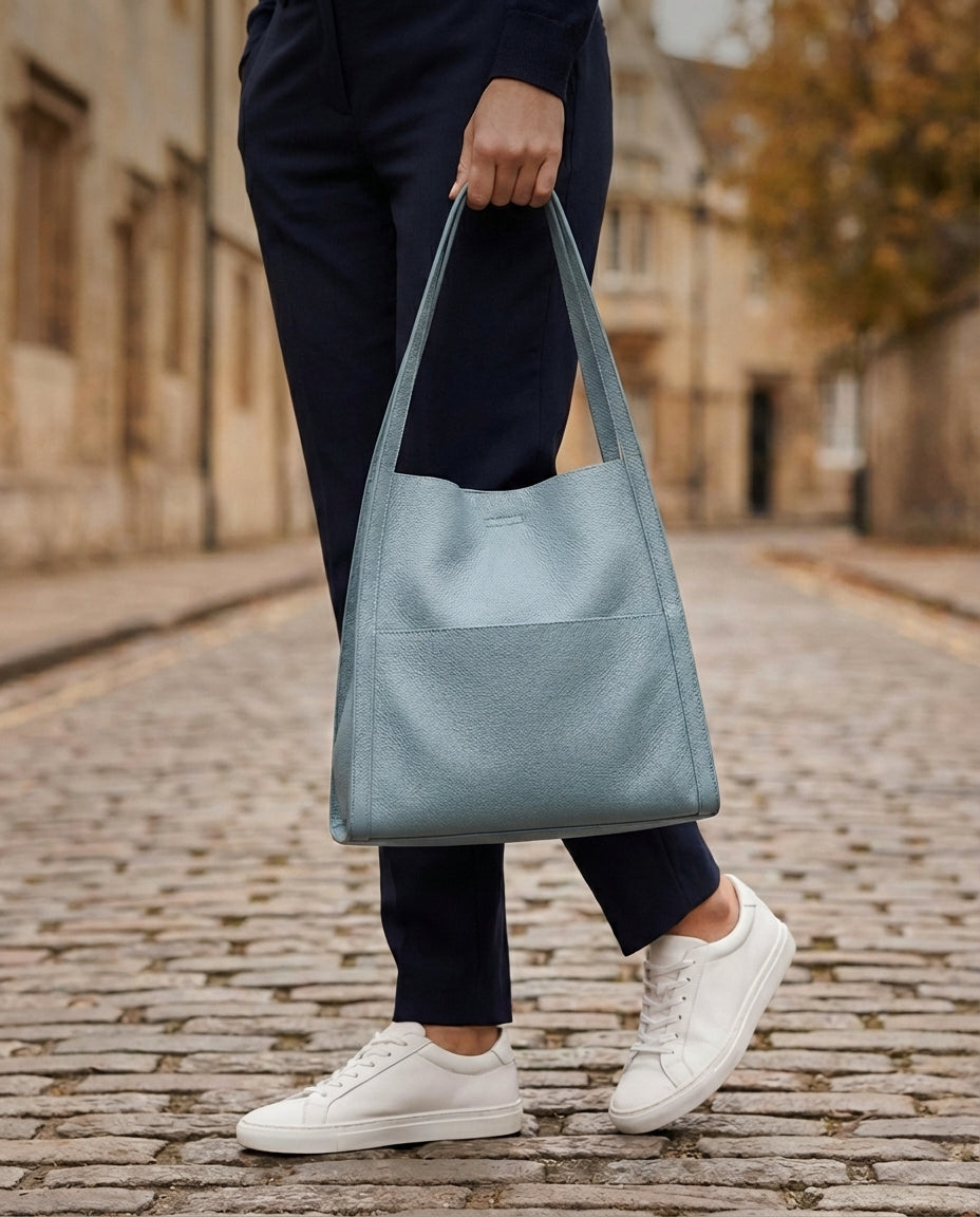 Person on a cobbled street holding the light blue leather tote by its straps, wearing navy trousers and white trainers. 6