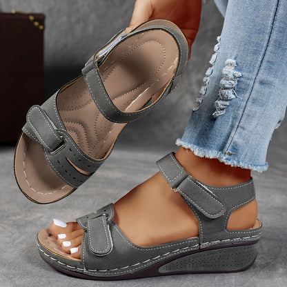 Grey wedge sandals with stitched straps, one on the foot and the other held up displaying the cushioned insole. 6
