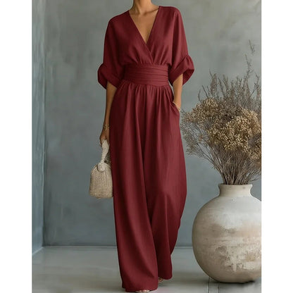 Burgundy version of the wide-leg jumpsuit with rolled half sleeves, worn by a model holding a woven bag beside a dried-flower vase. 2