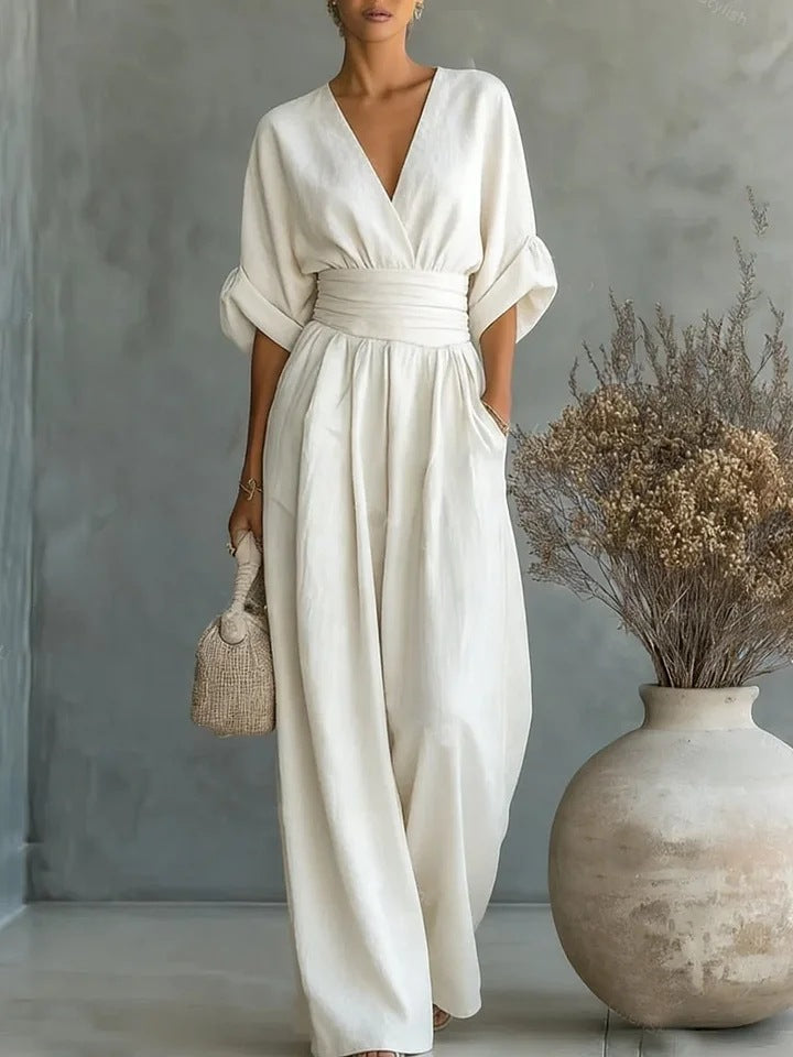 Cream wide-leg jumpsuit with V-neck and pleated waistband, full-length studio view with a woven bag and dried flowers in a vase. 9