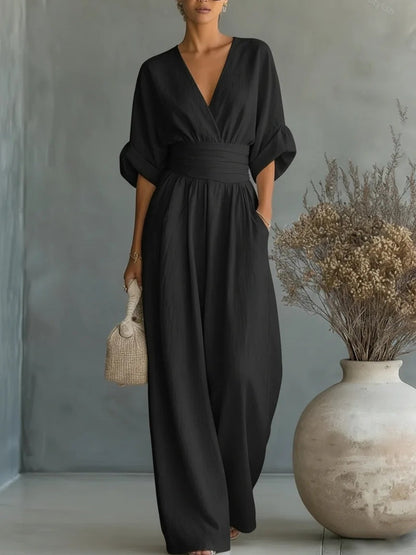Black version of the wide-leg jumpsuit with V-neckline and wide waistband, worn with a hand in the pocket in a studio setting. 3