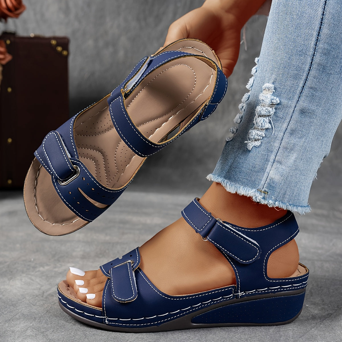 Navy blue wedge sandals with white stitching, one worn with ripped jeans and one lifted to show the insole. 8