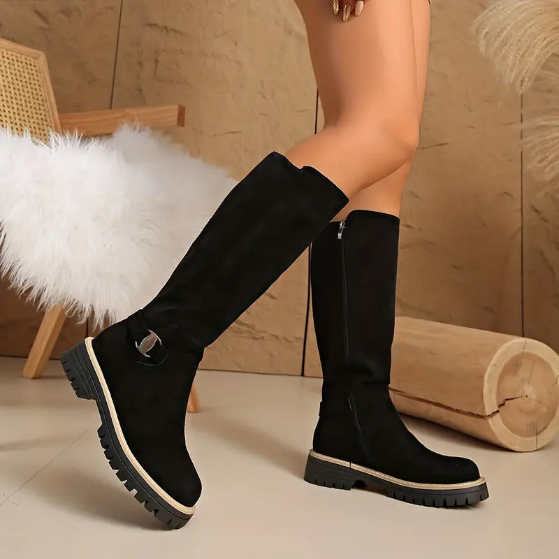 Black suede-look knee-high boots worn by a model, one foot angled to show the gold buckle strap and chunky lugged sole. 1