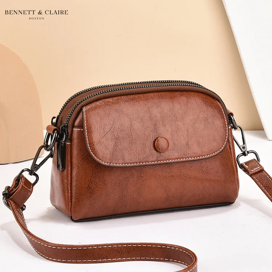 Tan-brown crossbody bag with stitched front flap, press stud and double zip top, shown against a peach backdrop. 1