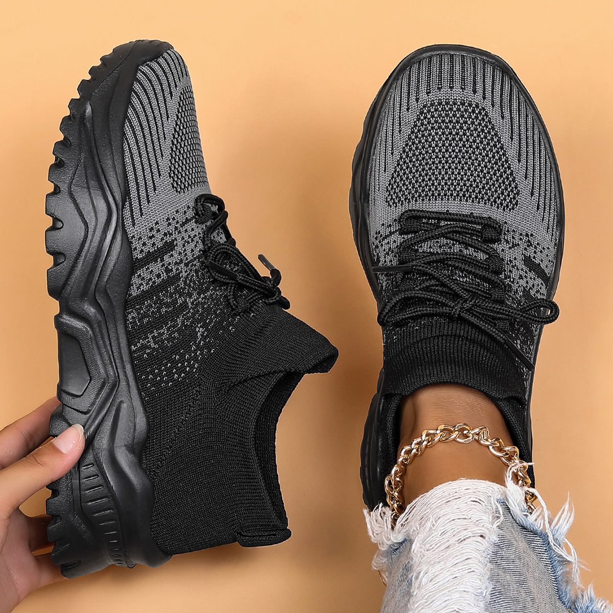 Top-down view of black and grey knit sock-style sneakers with chunky treaded soles, one held up and one worn with a gold anklet. 1