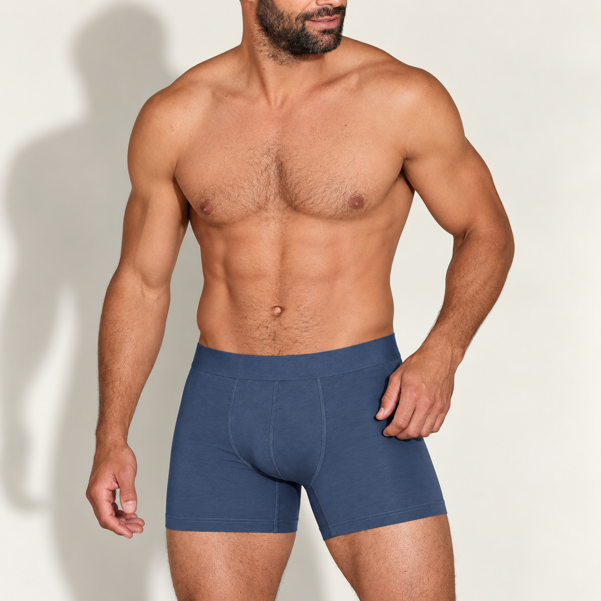 Bearded shirtless model standing at a slight angle, wearing mid-blue boxer briefs with a plain wide waistband and front seams. 1
