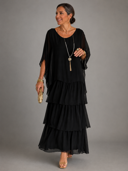 Woman wearing the black tiered chiffon maxi dress with a sheer cape overlay, gold clutch, long pendant necklace and gold heels, front view. 1