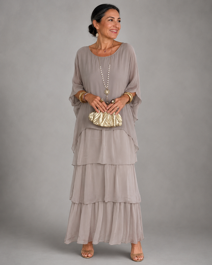 Woman wearing the taupe grey tiered chiffon maxi dress with a loose cape top, holding a gold ruched clutch. 3