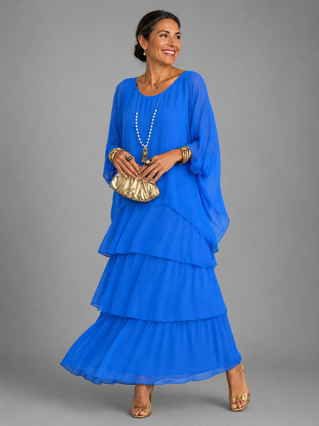 Woman wearing the bright royal blue tiered chiffon maxi dress with loose cape sleeves, holding a gold clutch and wearing gold sandals. 5