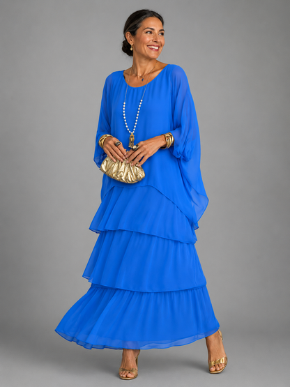 Woman wearing the bright royal blue tiered chiffon maxi dress with loose cape sleeves, holding a gold clutch and wearing gold sandals. 5