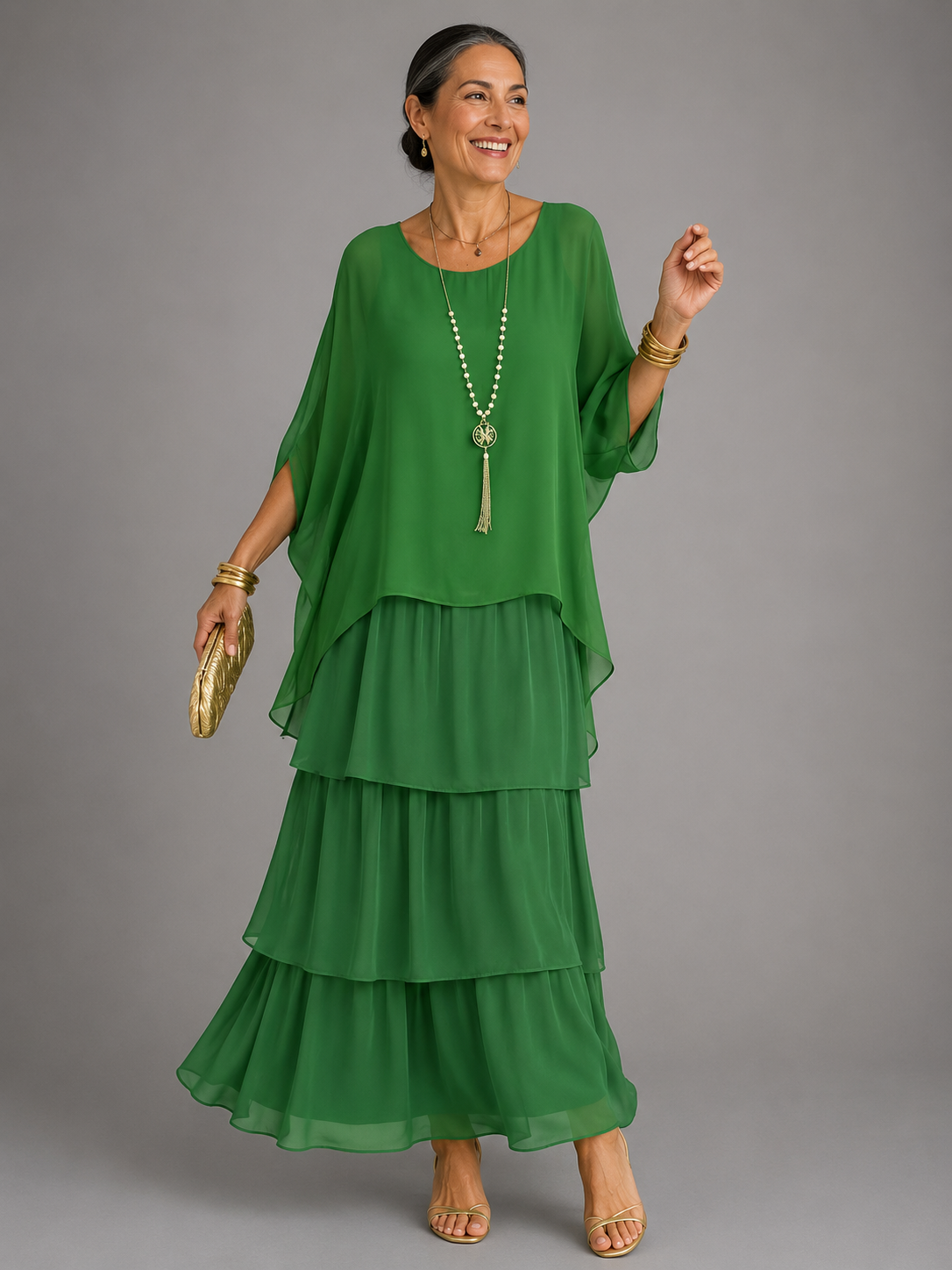 Woman wearing the emerald green tiered chiffon maxi dress with a sheer cape top, long tassel necklace and gold clutch in one hand. 7