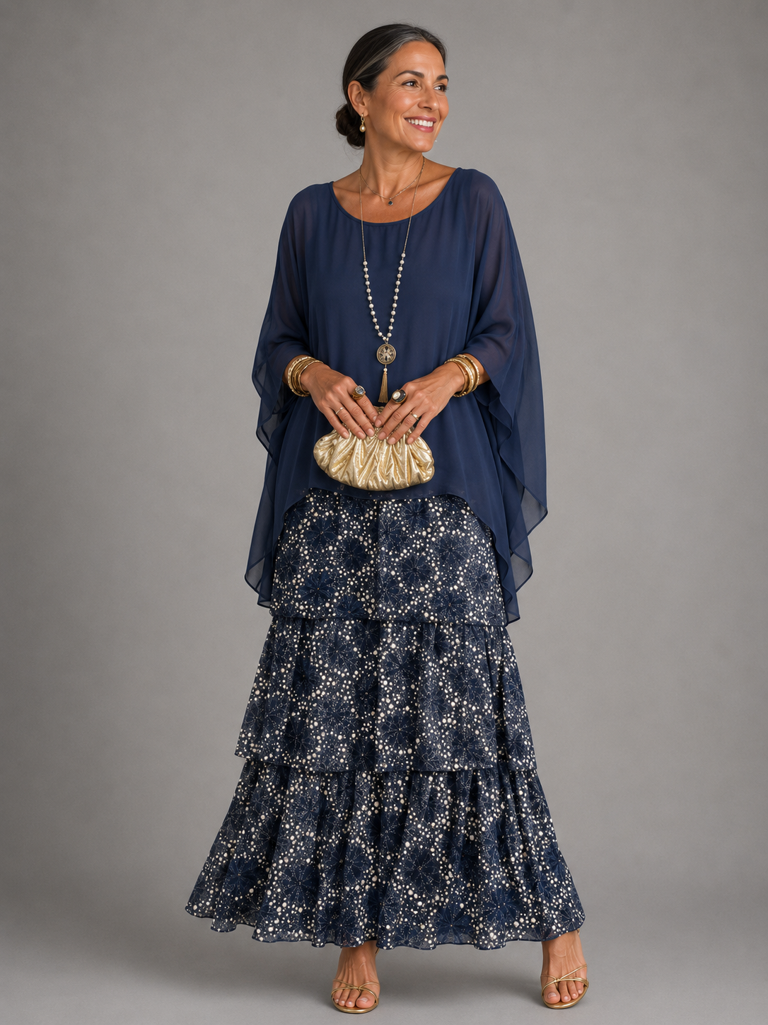 Woman wearing a navy cape top over a tiered maxi skirt with a white dotted floral pattern, gold clutch in hand. 11