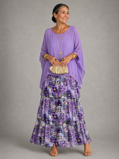 Woman in a lilac cape top over a tiered maxi skirt with a purple pansy floral print, holding a gold clutch. 10