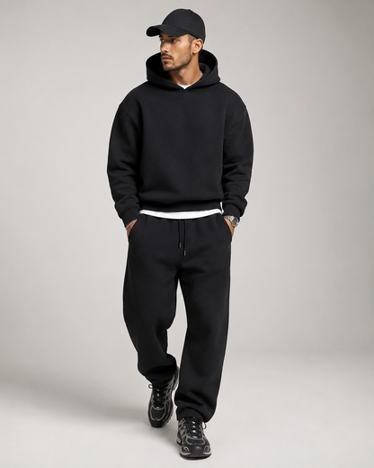 Model in the black hoodie and joggers walking, wearing a black cap, white layer and chunky black trainers, looking to one side. 7