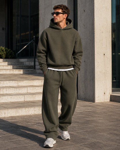 Model in the dark olive green hoodie and joggers outdoors in sunlight, standing by concrete pillars with hands in pockets. 16