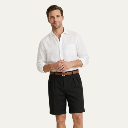 Man with clasped hands wearing black pleated turn-up shorts, a tan belt and a white linen shirt. 10