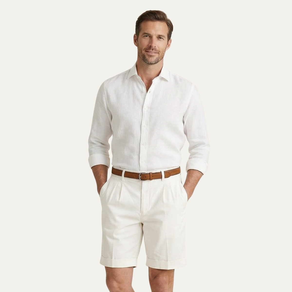 Studio shot of a man in cream pleated shorts with turn-up hems, white linen shirt and tan belt, hands in pockets. 6