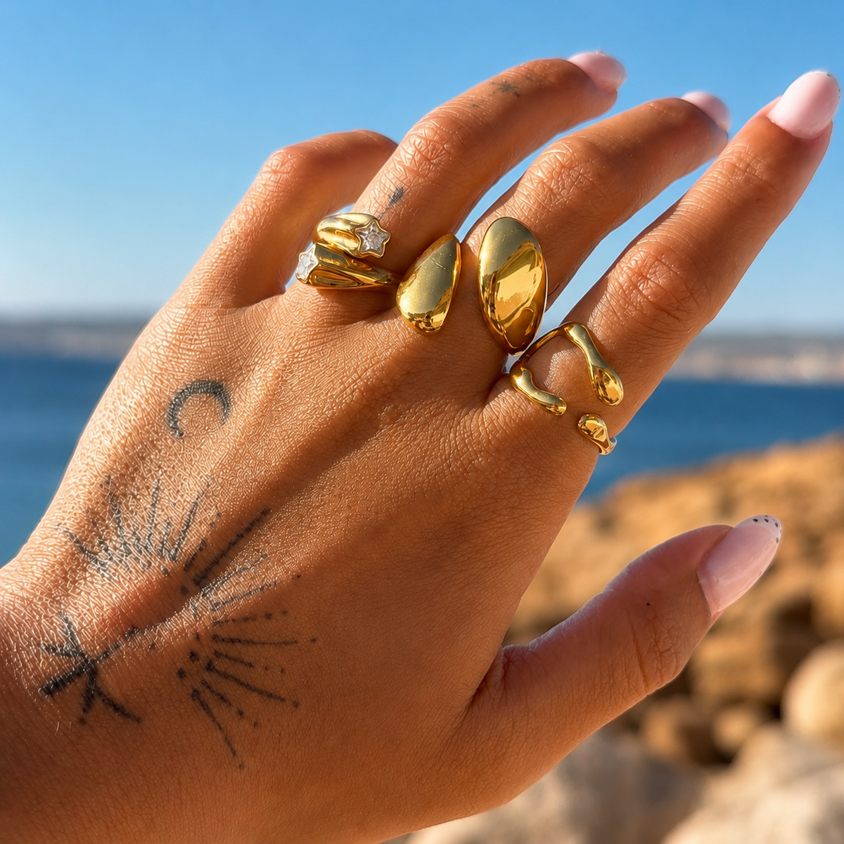 Hand with tattoos wearing several chunky gold-toned rings against a blue sea and clear sky, with the open ring on the middle finger. 3