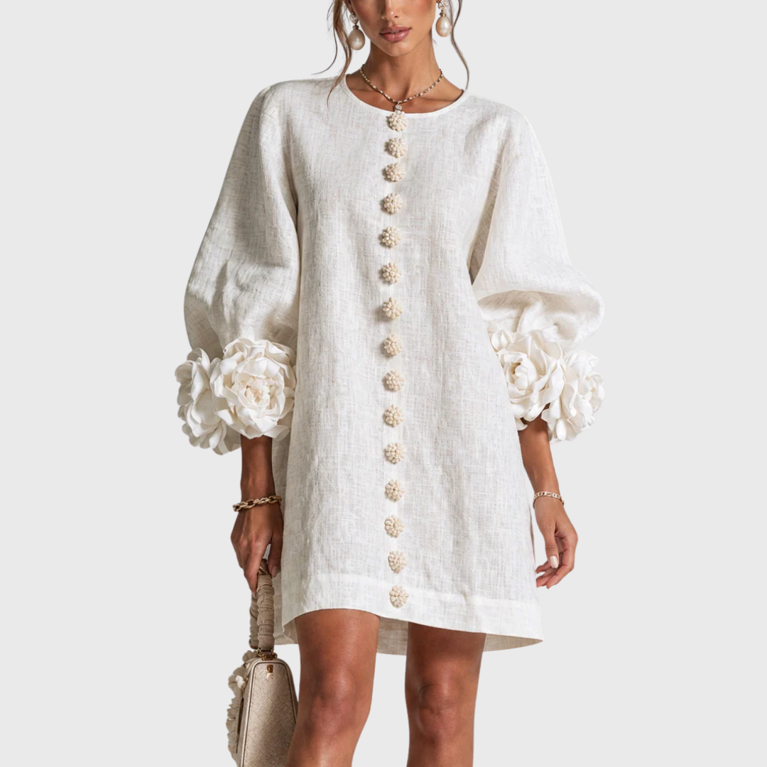 Front view of the white linen-look mini dress worn by a model, with flower buttons down the centre and rosette cuffs. 1