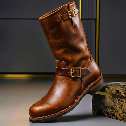 Tan-brown leather engineer boot with brass buckles on the shaft and ankle strap, shown on a grey studio backdrop. 1