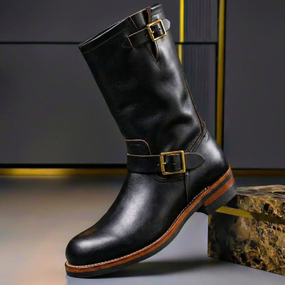 Black leather engineer boot with brass buckles on the shaft and ankle strap, tilted against a marble-effect block. 3